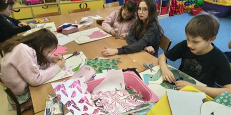 Powiększ grafikę: Grupa dzieci siedzi przy stole w świetlicy i wykleja postać kota. Na stole leżą pojemniki z kolorowymi kartkami, piankami, filcem, kredkami itp.