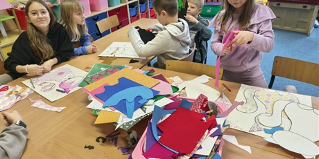 Powiększ grafikę: Grupa dzieci siedzi przy stole w świetlicy i wykleja postać kota. Na stole leżą pojemniki z kolorowymi kartkami, piankami, filcem, kredkami itp.