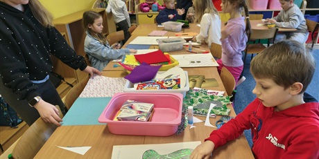 Powiększ grafikę: Grupa dzieci siedzi przy stole w świetlicy i wykleja postać kota. Na stole leżą pojemniki z kolorowymi kartkami, piankami, filcem, kredkami itp.