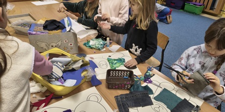 Powiększ grafikę: Grupa dziewczynek siedzi lub stoi przy stole w świetlicy. Dzieci wyklejają misie kolorowymi tkaninami, piankami itp. Na stole stoją kartony i kolorowe pojemniki z różnymi materiałami.