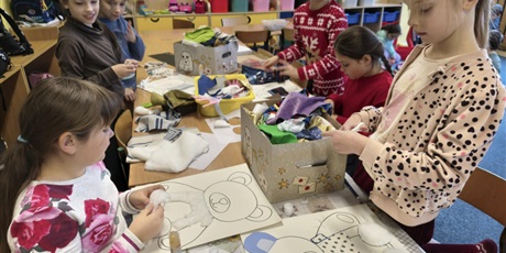 Powiększ grafikę: Grupa dziewczynek siedzi lub stoi przy stole w świetlicy. Dzieci wyklejają misie kolorowymi tkaninami, piankami itp. Na stole stoją kartony i kolorowe pojemniki z różnymi materiałami.