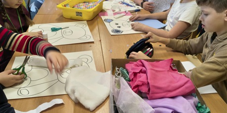 Powiększ grafikę: Grupa dzieci siedzi lub stoi przy stole w świetlicy. Dzieci wyklejają misie kolorowymi tkaninami, piankami itp. Na stole stoją kartony i kolorowe pojemniki z różnymi materiałami.