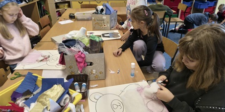 Powiększ grafikę: Grupa dziewczynek siedzi lub stoi przy stole w świetlicy. Dzieci wyklejają misie kolorowymi tkaninami, piankami itp. Na stole stoją kartony i kolorowe pojemniki z różnymi materiałami.