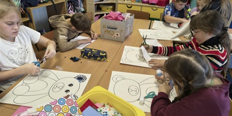 Powiększ grafikę: Grupa dzieci siedzi lub stoi przy stole w świetlicy. Dzieci wyklejają misie kolorowymi tkaninami, piankami itp. Na stole stoją kartony i kolorowe pojemniki z różnymi materiałami.