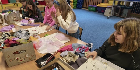 Powiększ grafikę: Grupa dzieci siedzi lub stoi przy stole w świetlicy. Dzieci wyklejają misie kolorowymi tkaninami, piankami itp. Na stole stoją kartony i kolorowe pojemniki z różnymi materiałami.