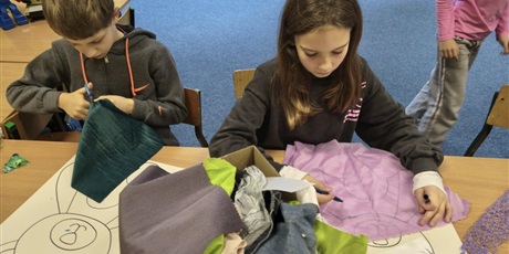 Powiększ grafikę: Dziewczynka i chłopiec siedzą przy stole w świetlicy. Oboje wyklejają misie kolorowymi tkaninami, piankami itp. Na stole stoi karton z różnymi, kolorowymi materiałami.