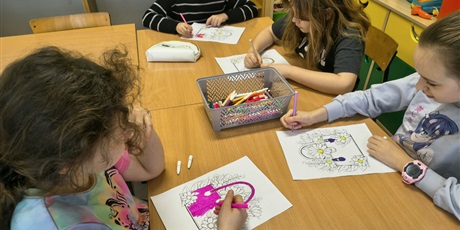 Powiększ grafikę: Grupa dziewczynek siedzi przy stole i koloruje kolorowankę relaksacyjną przedstawiającą damską torebkę. Na stole znajdują się kolorowe kredki i mazaki.
