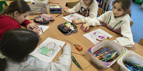 Powiększ grafikę: Grupa dziewczynek siedzi przy stole i koloruje kolorowankę relaksacyjną przedstawiającą damską torebkę. Na stole znajdują się kolorowe kredki i mazaki.