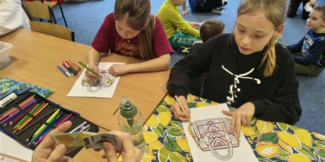 Powiększ grafikę: Dwie dziewczynki siedzą przy stole i koloruje kolorowankę relaksacyjną przedstawiającą damską torebkę. Na stole znajdują się kolorowe kredki i mazaki. Na drugim planie - na dywanie siedzą chłopcy i uk