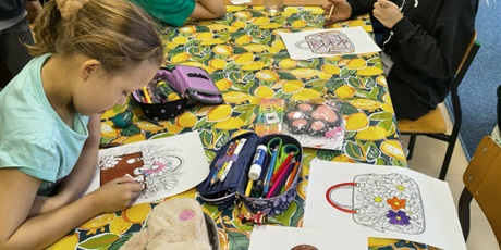 Powiększ grafikę: Grupa dziewczynek siedzi przy stole i koloruje kolorowankę relaksacyjną przedstawiającą damską torebkę. Na stole znajdują się kolorowe kredki i mazaki.