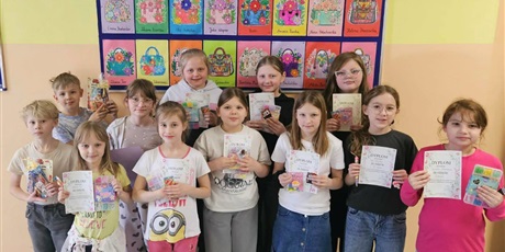 Powiększ grafikę: Grupa dzieci pozuje do zdjęcia na tle granatowej tablicy z kolorowymi pracami konkursowymi pt. "Projekt damskiej torebki". W rękach trzymają dyplomy i nagrody.