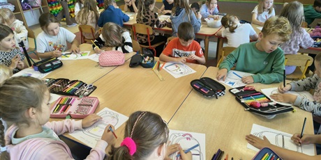 Powiększ grafikę: Grupa dzieci siedzi przy stole i koloruje projekt plecaka. Na stole leżą pojemniki i piórniki z kolorowymi kredkami.