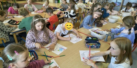 Powiększ grafikę: Grupa dzieci siedzi przy stole i koloruje projekt plecaka. Na stole leżą pojemniki i piórniki z kolorowymi kredkami.