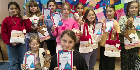 Powiększ grafikę: Grupa dziewczynek pozuje do zdjęcia.  W rękach trzymają dyplomy i nagrody. Za nimi znajduje się granatowa tablica, na której wiszą prace plastyczne, które przedstawiają kolorowe skarpety z motywami św