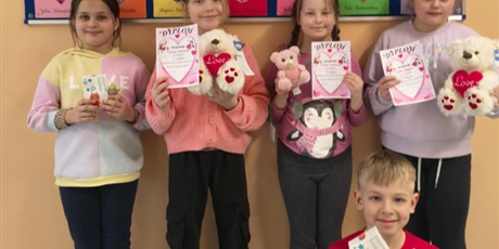 Powiększ grafikę: Grupa dzieci pozuje do zdjęcia. W rękach trzymają dyplomy i narody za udział w konkursie. Za nimi na ścianie wisi granatowa tablica z kolorowymi kartkami i sercami.