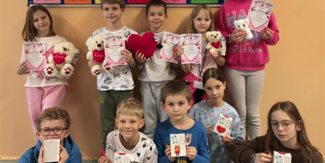 Powiększ grafikę: Grupa dzieci pozuje do zdjęcia. W rękach trzymają dyplomy i narody za udział w konkursie. Za nimi na ścianie wisi granatowa tablica z kolorowymi kartkami i sercami.