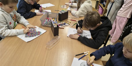 Powiększ grafikę: Grupa dzieci siedzi przy stole i koloruje kolorowankę relaksacyjną w kształcie serca. Na stole znajdują się mazaki i pojemniki z flamastrami.