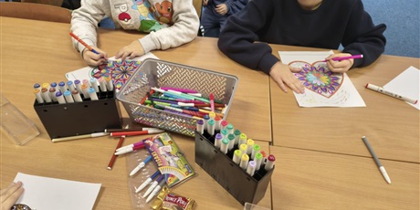 Powiększ grafikę: Dwóch chłopców siedzi przy stole i koloruje kolorowankę relaksacyjną w kształcie serca. Na stole znajdują się mazaki i pojemniki z flamastrami.