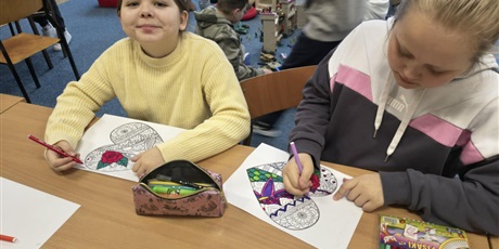 Powiększ grafikę: Dwie dziewczynki siedzą przy stole i kolorują kolorowankę relaksacyjną w kształcie serca. Na stole  znajdują się mazaki.