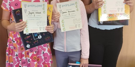 Powiększ grafikę: Na tle jasnobrązowej ściany stoją trzy dziewczynki, każda z promiennym uśmiechem na twarzy z dumą prezentują w dłoniach dyplomy oraz nagrody zdobyte w konkursie plastycznym. Ich radość i entuzjazm odd