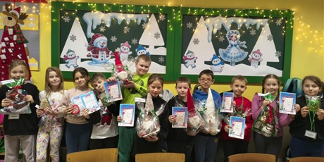 Powiększ grafikę: Grupa dzieci pozuje do zdjęcia. W rękach trzymają dyplomy i nagrody. W tle znajdują się dwie tablice ścienne z zimową dekoracją.