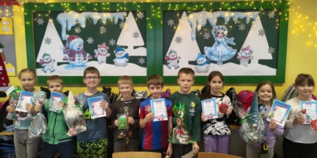 Powiększ grafikę: Grupa dzieci pozuje do zdjęcia. W rękach trzymają dyplomy i nagrody. W tle znajdują się dwie tablice ścienne z zimową dekoracją.