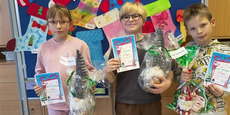 Powiększ grafikę: Troje dzieci pozuje do zdjęcia. W rękach trzymają dyplomy i nagrody. W tle znajduje się granatowa tablica, na której wiszą świąteczne skarpety.