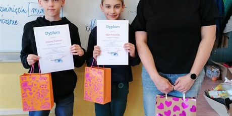 Powiększ grafikę: Nauczycielka wraz z dwoma uczniami stoją w klasie, w tle tablica. Prezentują dyplomy. 