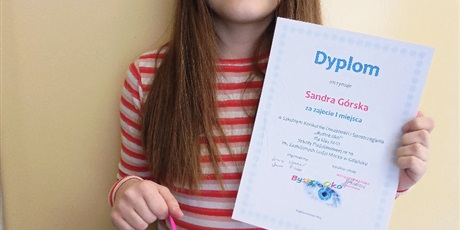 Powiększ grafikę: Dziewczynka stoi na tle ściany. W rękach trzyma dyplom i nagrodę. 