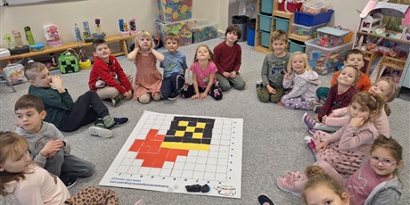 Powiększ grafikę: Grupa zerówkowiczów siedzi w kole na dywanie. Przed nimi leży efekt kodowania, czyli rysunek złożony z kwadratów o różnych kolorach. Wszyscy są bardzo dumni z wykonanej pracy.