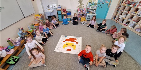 Powiększ grafikę: Grupa zerówkowiczów siedzi w kole na dywanie. Przed nimi leży efekt kodowania, czyli rysunek twarzy Mikołaja złożony z kwadratów o różnych kolorach. Wszyscy są bardzo dumni z wykonanej pracy.