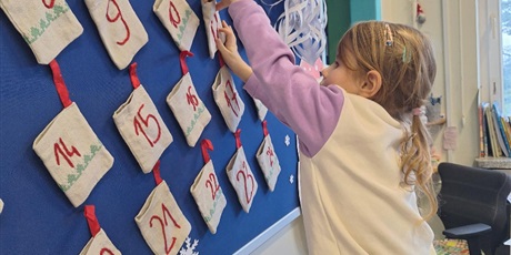 Powiększ grafikę: Dziewczynka otwiera okienku w kalendarzu adwentowym. Na tablicy zwieszone są woreczki z datami. 