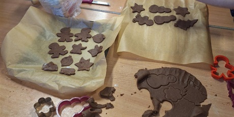 Powiększ grafikę: Dzieci przygotowują pierniki. Z rozwałkowanego ciasta wykrawają foremkami wzory świątecznych pierniczków i przekładają je na blaszkę do pieczenia.