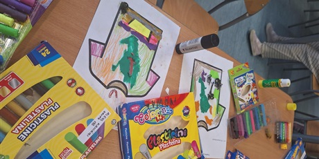 Powiększ grafikę: Na ławce szkolnej znajdują się rysunki oraz materiały plastyczne. Widać projekt koszulki piłkarskiej.