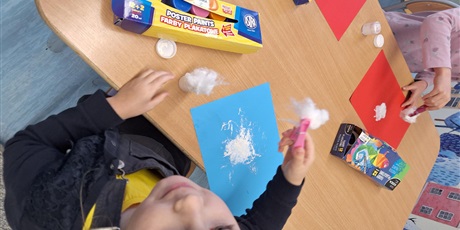 Powiększ grafikę: Na zdjęciu widać dziecko, które wykonuje pracę plastyczną. Na kolorowej kartce, za pomocą waty trzymanej w spinaczu do prania, dziecko robi kleksy z białej farby.