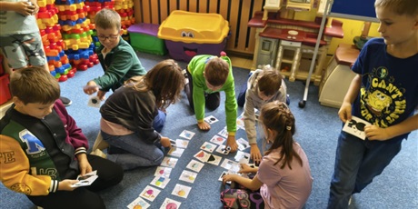 Powiększ grafikę: Grupa dzieci pochyla się nad kartami przedstawiającymi kolorowe kropki i ich cienie.    Dwóch chłopców stoi. W rękach dzieci trzymają karty. 