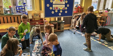 Powiększ grafikę: Dwie grupy dzieci pochylają się nad kartami przedstawiającymi kolorowe kropki i ich cienie.  W rękach dzieci trzymają karty i układają je na dywanie. Jeden chłopiec stoi.