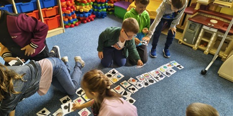 Powiększ grafikę: Grupa dzieci pochyla się nad kartami przedstawiającymi kolorowe kropki i ich cienie.  W rękach dzieci trzymają karty i układają je na dywanie.