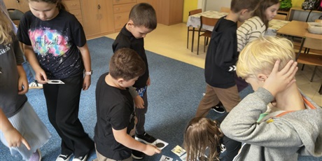 Powiększ grafikę: Grupa dzieci pochyla się nad kartami przedstawiającymi kolorowe kropki i ich cienie.  W rękach dzieci trzymają karty i układają je na dywanie.