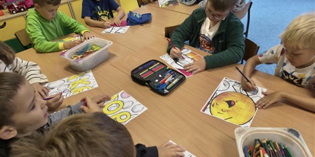 Powiększ grafikę: Chłopcy siedzą przy stołach i kolorują kartki z kropkami. Na stole znajdują się pojemniki z kredkami i piórniki.