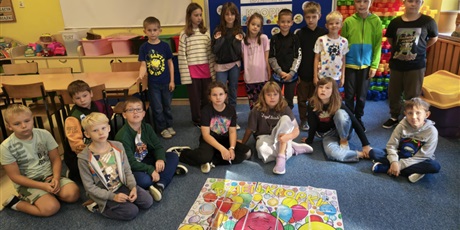 Powiększ grafikę: Grupa dzieci pozuje do zdjęcia. Dzieci w pierwszym rzędzi siedzą na dywanie. przed nimi leży plakat. W drugim rzędzie dzieci stoją. Za nimi znajduje się granatowa tablica z kolorową dekoracją.