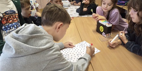 Powiększ grafikę: Dzieci siedzą lub stoją przy stole. Na stole znajduje się kartka z kropkami, na której dzieci rozgrywają pojedynek pt. "Zgarnij kropki".