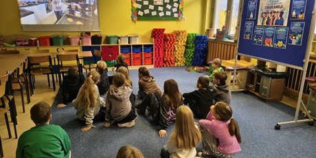 Powiększ grafikę: Grupa dzieci na dywanie w świetlicy tyłem do obiektywu. Dzieci oglądają film wyświetlany na ściennym ekranie.