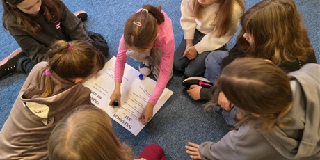 Powiększ grafikę: Grupa dziewczynek siedzi na dywanie w świetlicy. Pochylają się nad plakatem, na który przyklejają karteczki z przykładami tolerancji.