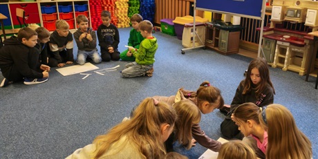 Powiększ grafikę: Dwie grupy dziewczynek i chłopców siedzą na dywanie w świetlicy.  Dzieci pochylają się nad plakatem, na który przyklejają karteczki z przykładami tolerancji.