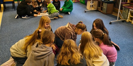 Powiększ grafikę: Dwie grupy dziewczynek i chłopców siedzą na dywanie w świetlicy.  Dzieci pochylają się nad plakatem, na który przyklejają karteczki z przykładami tolerancji.