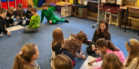 Powiększ grafikę: Dwie grupy dziewczynek i chłopców siedzą na dywanie w świetlicy.  Dzieci pochylają się nad plakatem, na który przyklejają karteczki z przykładami tolerancji.