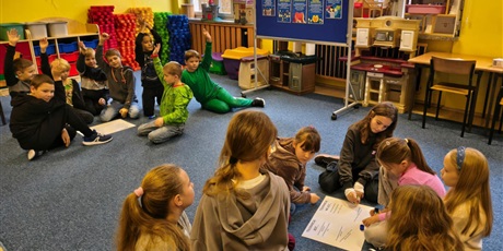 Powiększ grafikę: Dwie grupy dziewczynek i chłopców siedzą na dywanie w świetlicy.  Dzieci pochylają się nad plakatem, na który przyklejają karteczki z przykładami tolerancji.