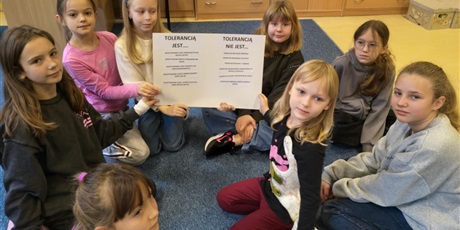 Powiększ grafikę: Grupa dziewczynek siedzi na dywanie w świetlicy. Dwie dziewczynki trzymają w rękach plakat uniesiony do góry, który wspólnie wykonały.