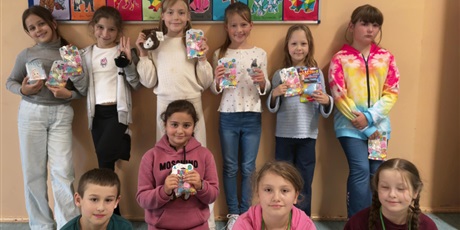 Powiększ grafikę: Grupa dzieci pozuje do zdjęcia. W rękach trzymają upominki otrzymane w ramach konkursu "Zwierzęta w geometrii". Na drugim planie znajduje się tablica z powieszonymi kolorowankami zwierząt.